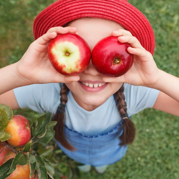 A little girl in a blue apron put two apples to her eyes on a background of grass