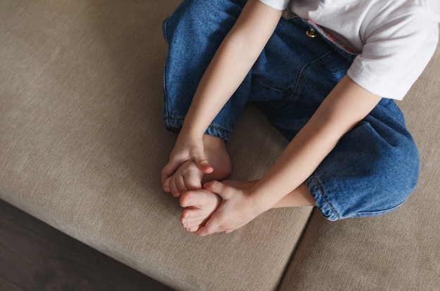 A little girl in a blue denim jumpsuit is sitting on the couch with her arms wrapped around her legs. Close-up