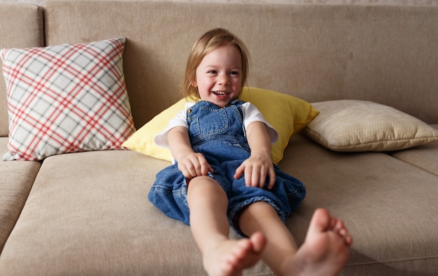 A little girl in a blue denim jumpsuit lies at home on the sofa among the colorful pillows and smiles cheerfully