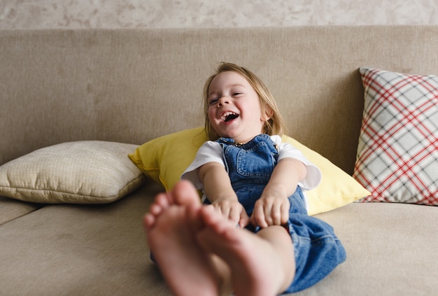 A little girl in a blue denim jumpsuit lies at home on the sofa among the colorful pillows and smiles cheerfully