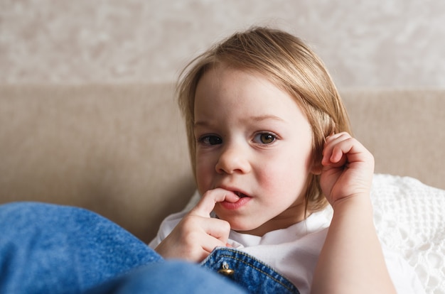 A little girl in a blue denim jumpsuit sits on the couch and bites her finger nails. Close-up