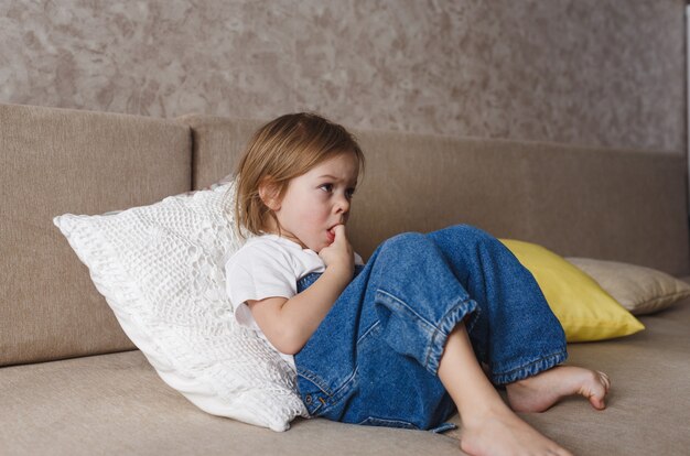 A little girl in a blue denim jumpsuit sits on the couch and bites her finger nails