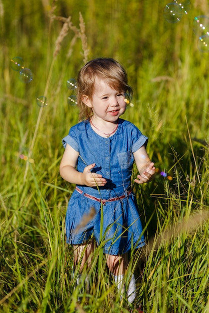 Little girl in blue dress trying to catch soap bubbles in summer park