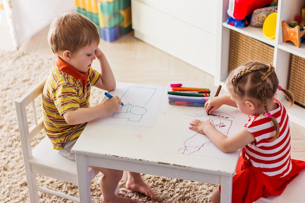 Little girl and boy at home sit at the table and draw with markers