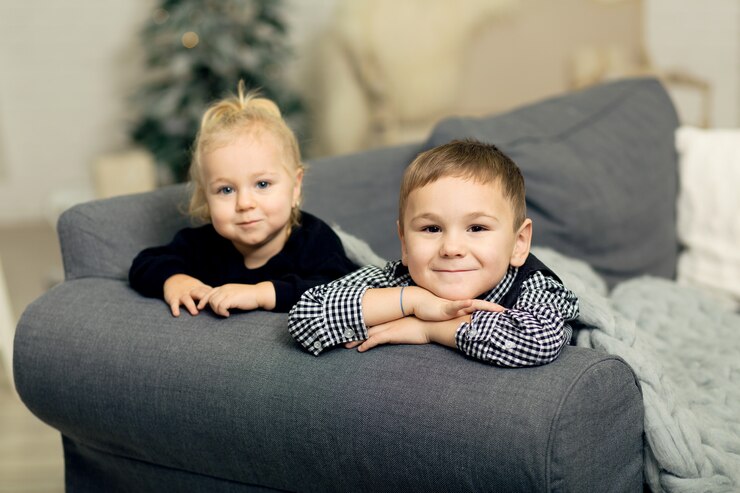 Photo little girl and boy lying t on the couch covered with a gray knitted blanket and smiling. brother and sister grimacing and fooling around on the  of new year's decor and christmas tree.