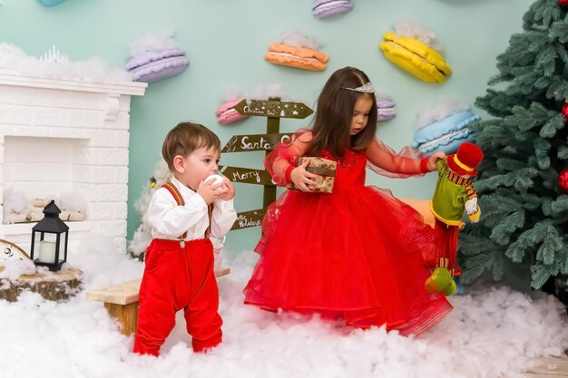 A little girl and a boy in red clothes have fun in the artificial snow by the fireplace in a studio