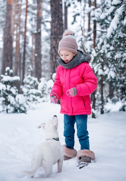 Little girl in a bright jacket plays in the winter snowy forest with her dog jack russell terrier