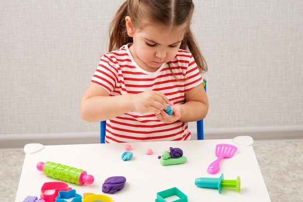 밝은 줄무늬 티셔츠를 입은 어린 소녀가 흰색 탁자에서 달팽이를 만드는 플라스티신을 가지고 노는 가정 교육 게임 개념
