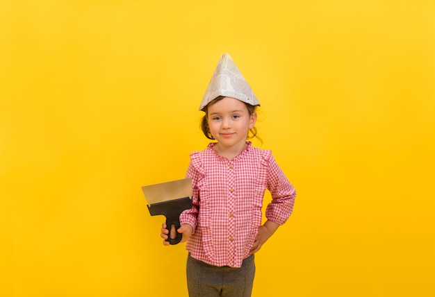 Un costruttore della bambina in un cappello di carta con una cazzuola di metallo su un giallo isolato