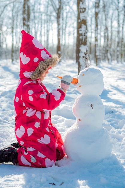 Little girl building snowman in snowy park Active outdoors leisure with family with children in winter Kid during stroll in a snowy winter park