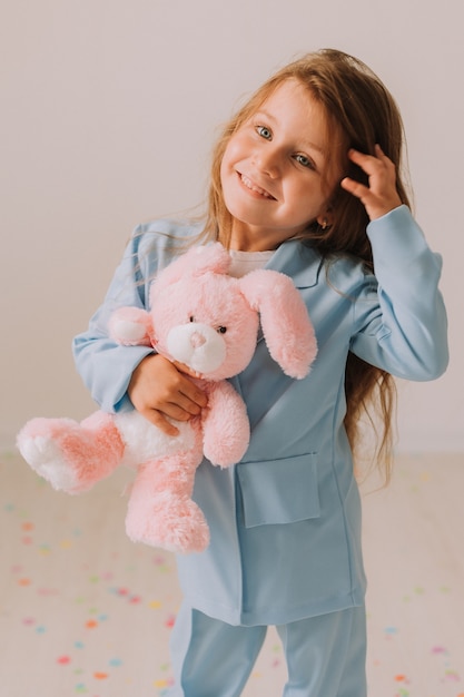 Little girl in a business blue pantsuit with a toy rabbit in her hands stands on a gray background