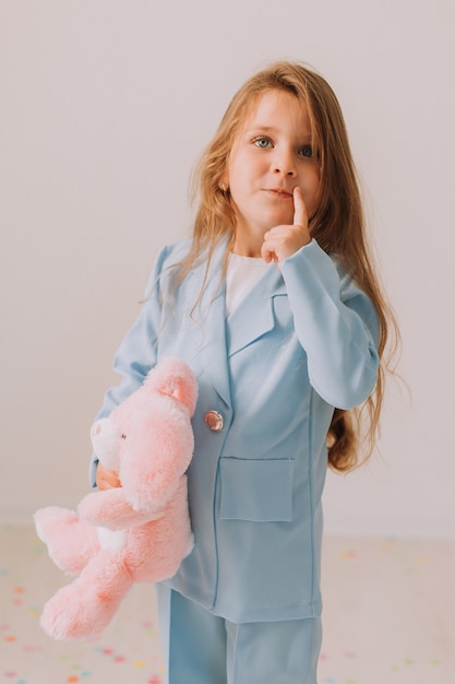 Little girl in a business blue pantsuit with a toy rabbit in her hands stands on a gray background