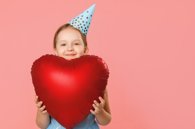 Маленькая девочка в кепке держит большой воздушный шар в форме сердца.