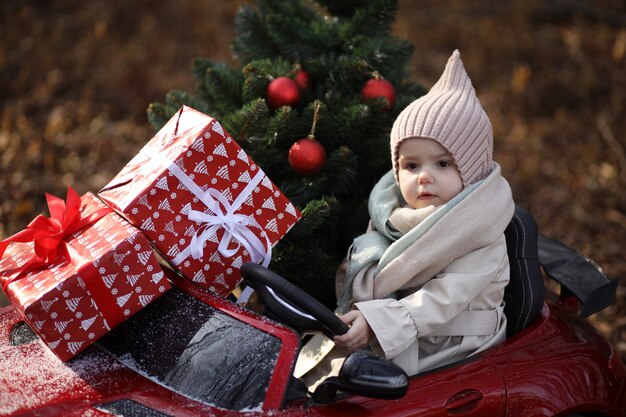 小さな女の子が赤い車でクリスマスツリーと贈り物を運ぶ