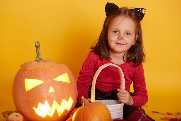 little girl in cat mask sitting and posing with terrifying carved pumpkin