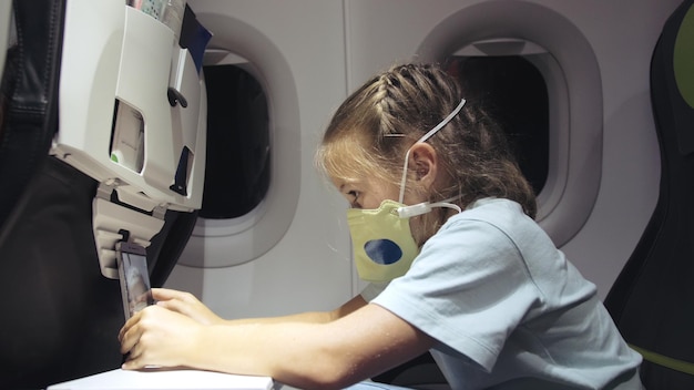 Little girl caucasian at plane with wearing protective mask Child baby tourist at aircraft with respirator play at smartphone mobile Covid19