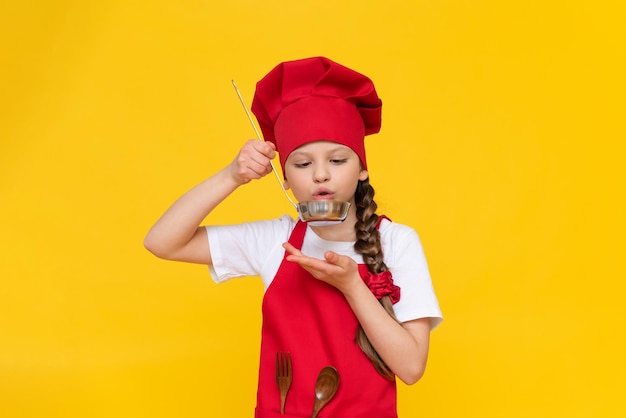 シェフの衣装を着た少女が料理を学んでいる 子供たちによる料理 赤いエプロンと黄色の孤立した背景に帽子をかぶった子供
