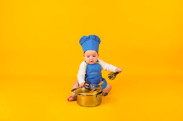 A little girl in a chef costume sits with a metal pot on a yellow wall with space for text