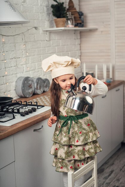 Little girl in a chef's cap examines a kettle in the kitchen