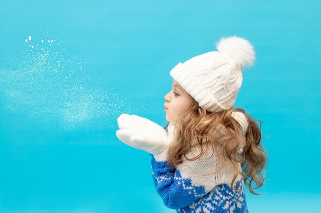 A little girl child on a blue background in a winter hat and sweater blows snow off her mittened hands, space for text