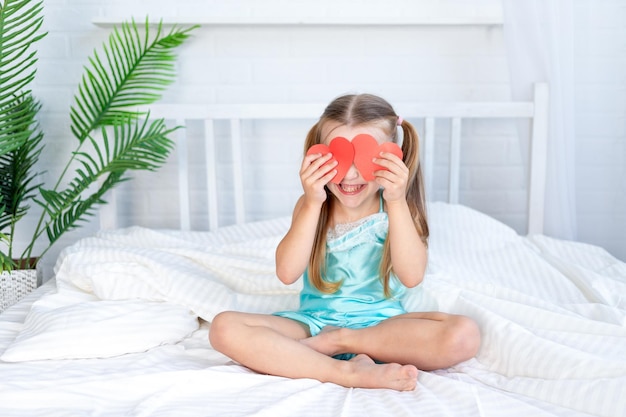 Little girl child closed her eyes with hearts sitting at home on the bed on a white cotton bed and smiling sweetly the concept of valentine's day