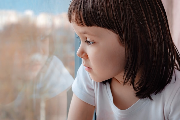 Una bambina a casa che guarda speranzoso attraverso il vetro della finestra.