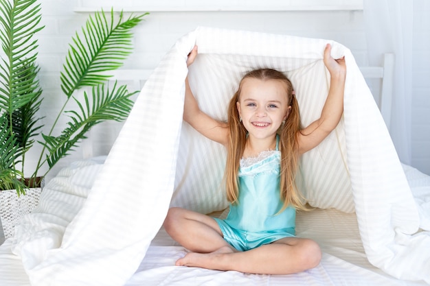 A little girl child is playing under the blanket waking up in the morning at home on the bed on a white cotton bed and smiling sweetly