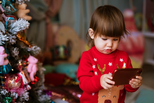 Little girl child playing mobile phone in red christmas sweater next to festive tree children and sm...