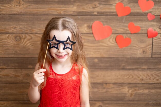 A little girl child in a red dress holds a mask glasses in the form of stars on a stick on a dark brown wooden background with red hearts the concept of Valentine's day a place for text