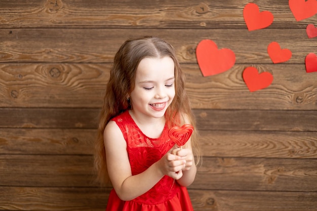 A little girl child in a red dress licks with her tongue a large lollipop in the form of a red heart on a dark brown wooden background with red hearts the concept of Valentines day a place for text