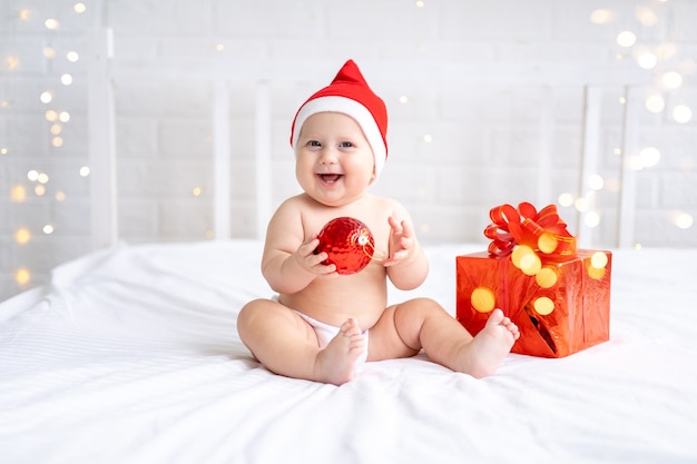 Una bambina con un cappello rosso da babbo natale è seduta su un letto bianco con scatole regalo su sfondo bianco