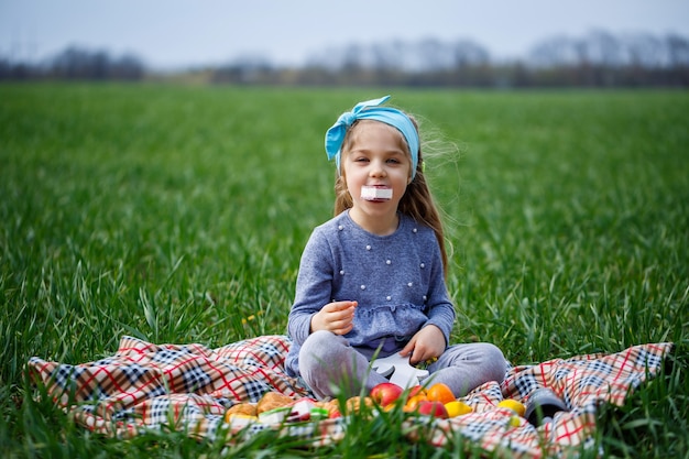 La bambina si siede sul copriletto e mangia biscotti e marmellata, erba verde nel campo, tempo primaverile soleggiato, sorriso e gioia del bambino, cielo blu con nuvole