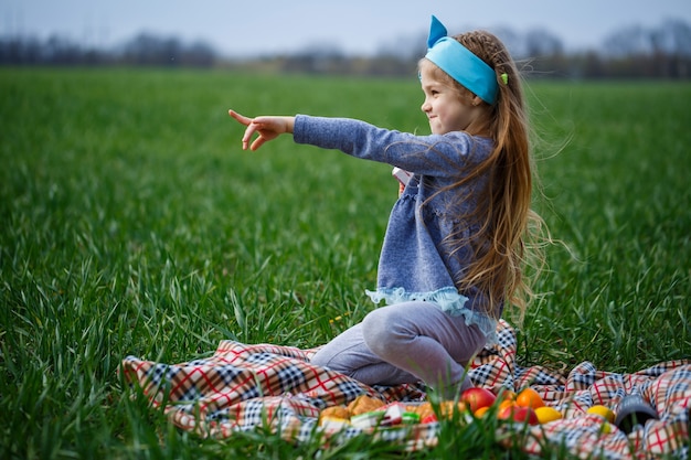 Little girl child sits on the bedspread and eats cookies and marmalade, green grass in the field, sunny spring weather, smile and joy of the child, blue sky with clouds