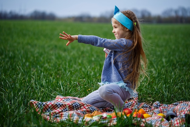 Little girl child sits on the bedspread and eats cookies and marmalade, green grass in the field, sunny spring weather, smile and joy of the child, blue sky with clouds