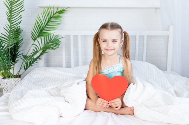 Little girl child with a big red heart in her hands sitting at home on the bed on a white cotton bed and smiling sweetly, valentine's day concept