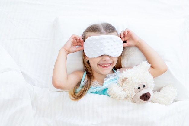 A little girl child with a sleep mask woke up in the morning at home on a bed on a white cotton bed with a teddy bear toy and a cute smile