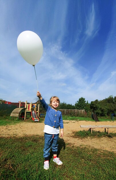 Фото Маленькая девочка с белым воздушным шаром в протянутой руке на улице на городской детской площадке