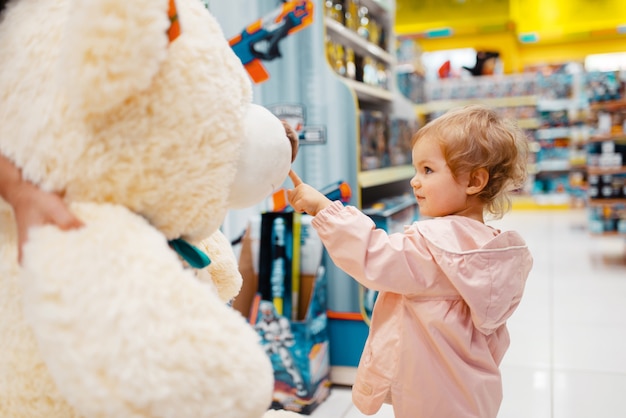 Little girl choosing big teddy bear in kids store, side view.