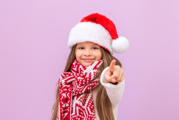 외딴 배경에 있는 크리스마스 의상을 입은 어린 소녀는 매우 행복해하며 손가락을 앞으로 가리킵니다. 아이는 크리스마스를 즐기고 있습니다.