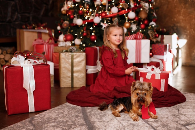 크리스마스 드레스를 입은 어린 소녀와 크리스마스 트리 근처의 집에 있는 강아지. 메리 크리스마스 인사