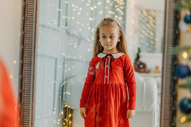 A little girl on christmas eve in festive clothes stands near the mirror and looks at her reflection. the child looks at himself in the mirror before the new year