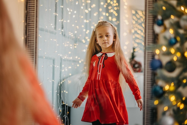 Маленькая девочка в канун Рождества в праздничной одежде стоит возле зеркала и смотрит на свое отражение. Ребенок смотрит на себя в зеркало перед Новым годом.