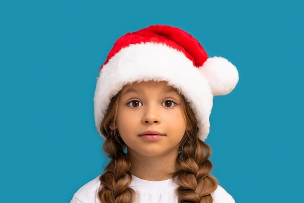 A little girl in a Christmas hat poses on a blue wall.