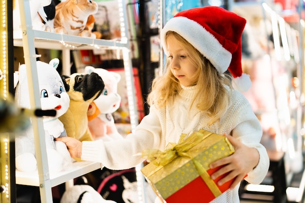 A little girl in a christmas hat with a new years gift in her hands at a showcase with toys children