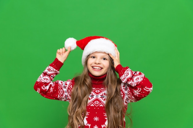 A little girl in a Christmas sweater plays with a Santa Claus hat in anticipation of the holiday on a green isolated background