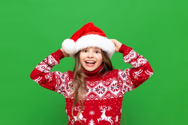 A little girl in a Christmas sweater plays with a Santa Claus hat in anticipation of the holiday on a green isolated background