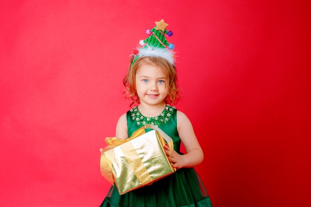 Little girl in a Christmas tree costume holds a gift on a red background new year