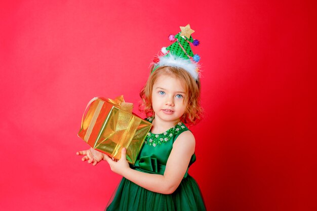 Little girl in a Christmas tree costume holds a gift on a red background new year