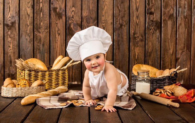 Little girl in a cook costume among a baking bread rolls of flour confectionery on a brown wooden floor sits on a wooden background background