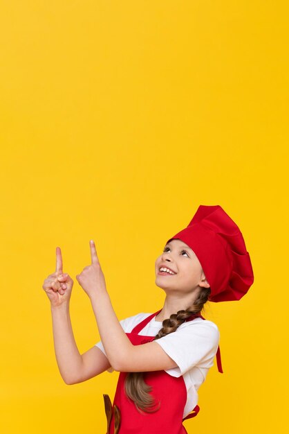 A little girl cook points her hands at the advertisement and smiles the concept of a child cooking at home a beautiful attractive girl on a yellow isolated background in a cook costume